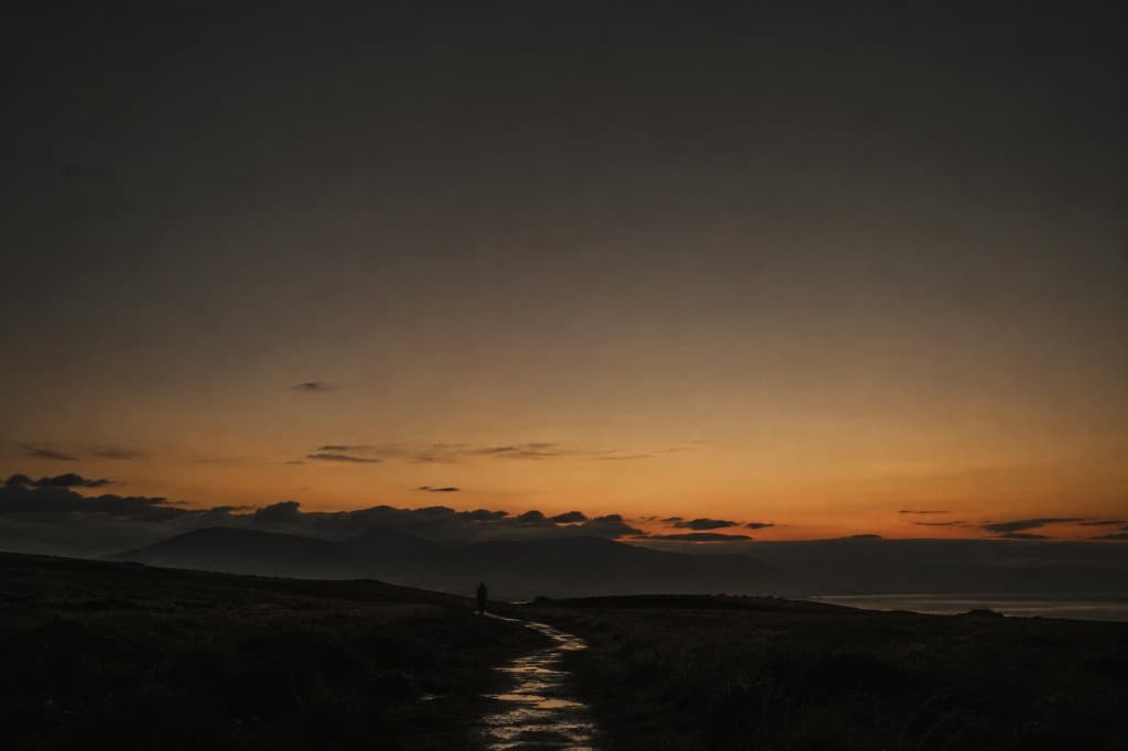 Silhouette on a wet path toward a low glowing horizon under a wide dark sky, reflective mood.